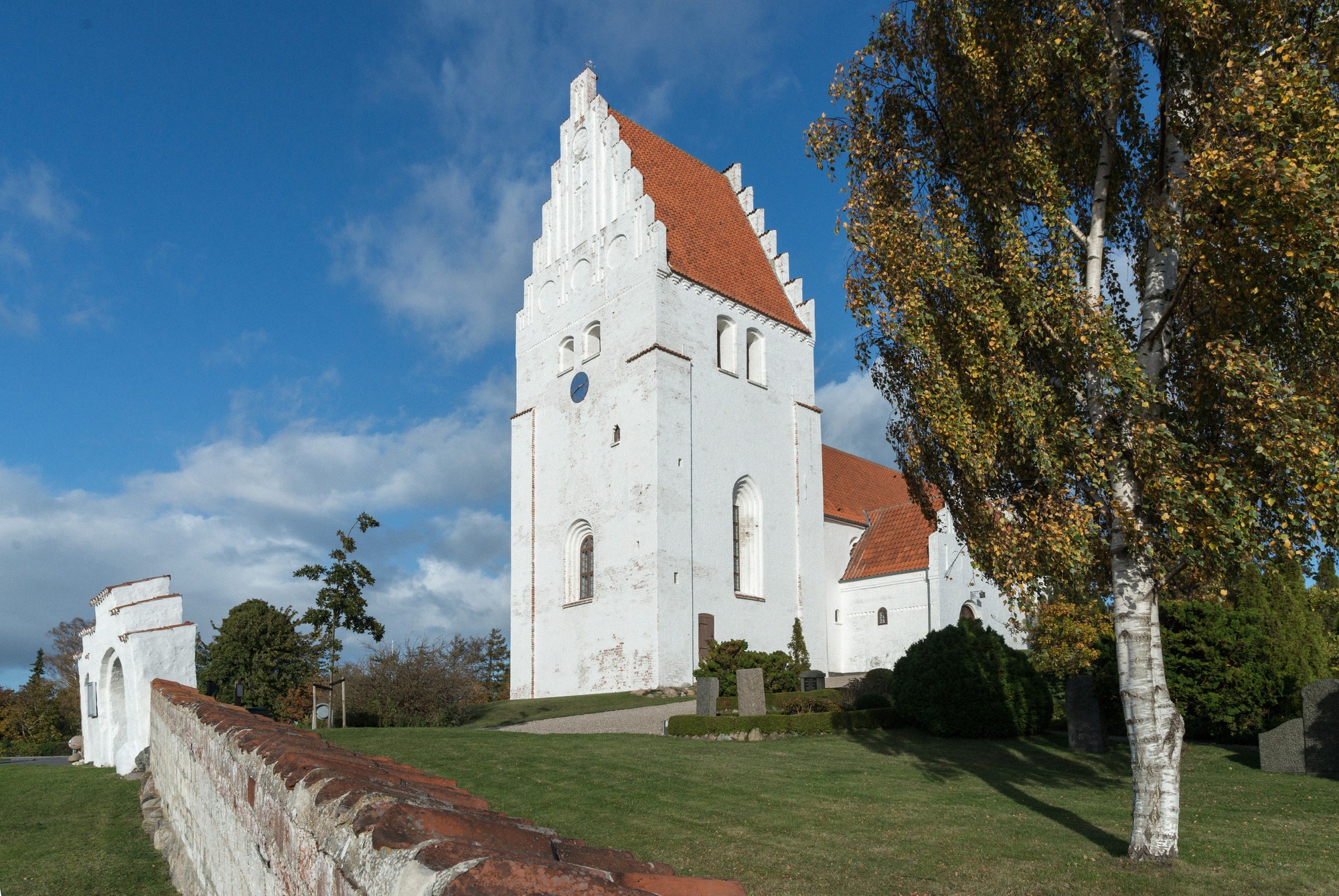 Die Elmelunde Kirke mit ihrem markanten weißen Turm und rotem Ziegeldach liegt idyllisch in grüner Landschaft.